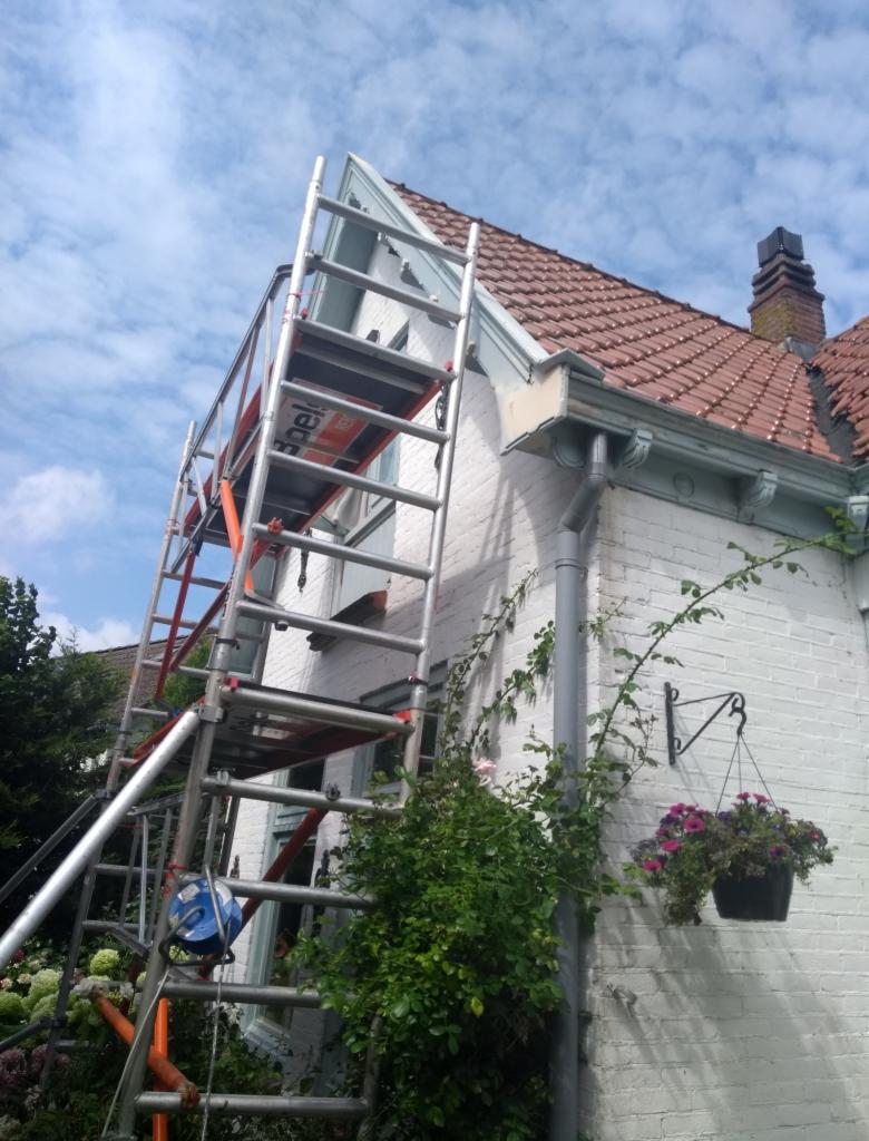 Rozstawiony aluminiowy ruszt przy białej, ceglanej ścianie domu z czerwonym dachem, widoczne rynny, wisząca doniczka z kwiatami i fragmenty zieleni.