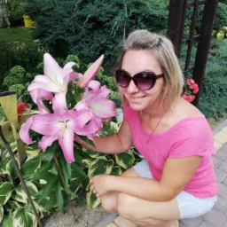 Uśmiechnięta kobieta w okularach kuca obok okazałych, różowych lilii w ogrodzie z bujną roślinnością w tle. Widoczne kostki brukowe i drewniana konstrukcja.