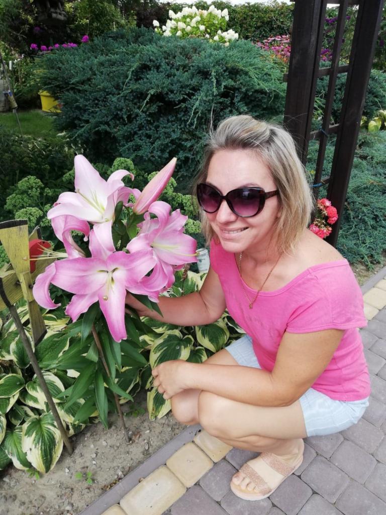 Uśmiechnięta kobieta w okularach kuca obok okazałych, różowych lilii w ogrodzie z bujną roślinnością w tle. Widoczne kostki brukowe i drewniana konstrukcja.