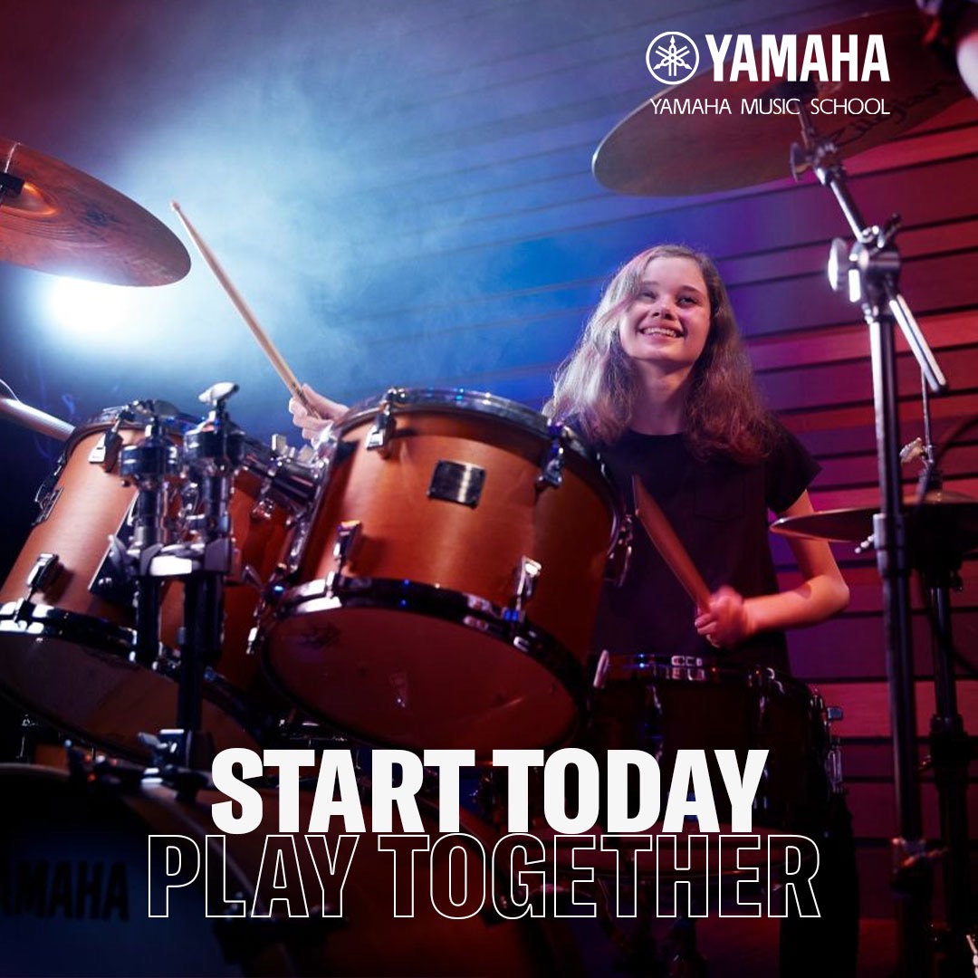 Uśmiechnięta dziewczyna gra na perkusji Yamaha w szkole muzycznej, oświetlona niebiesko-czerwonym światłem. Widoczne pałeczki i logo Yamaha Music School.