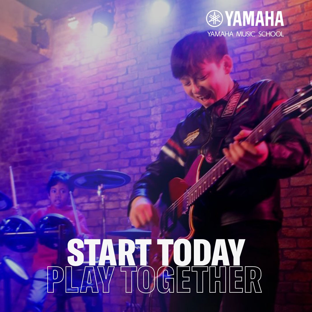 Uśmiechnięty chłopak gra na czerwonej gitarze elektrycznej, w tle perkusja i drugi chłopak. Logo Yamaha Music School w rogu. Ceglana ściana w tle.