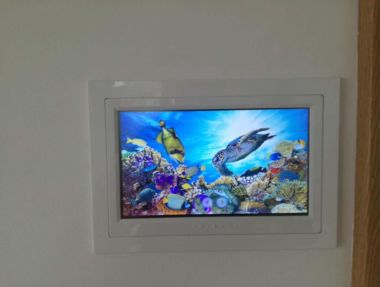 Panel dotykowy w białej ramce, wyświetlający animację podwodnego świata z żółwiem, rybami i rafą koralową, zamontowany na białej ścianie obok drewnianej framugi.