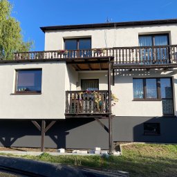 TERMOKOMPLEX MADEX Amadeusz Golik - Dwukondygnacyjny dom jednorodzinny z balkonami w Bestwince, otynkowany na biało z ciemnoszarą dolną partią elewacji, drewniane balustrady balkonów z donicami kwiatów, błękitne niebo w tle.