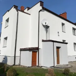 Trzykondygnacyjny dom jednorodzinny z białą elewacją, brązowymi elementami wykończeniowymi i anteną satelitarną, sfotografowany w słoneczny dzień.