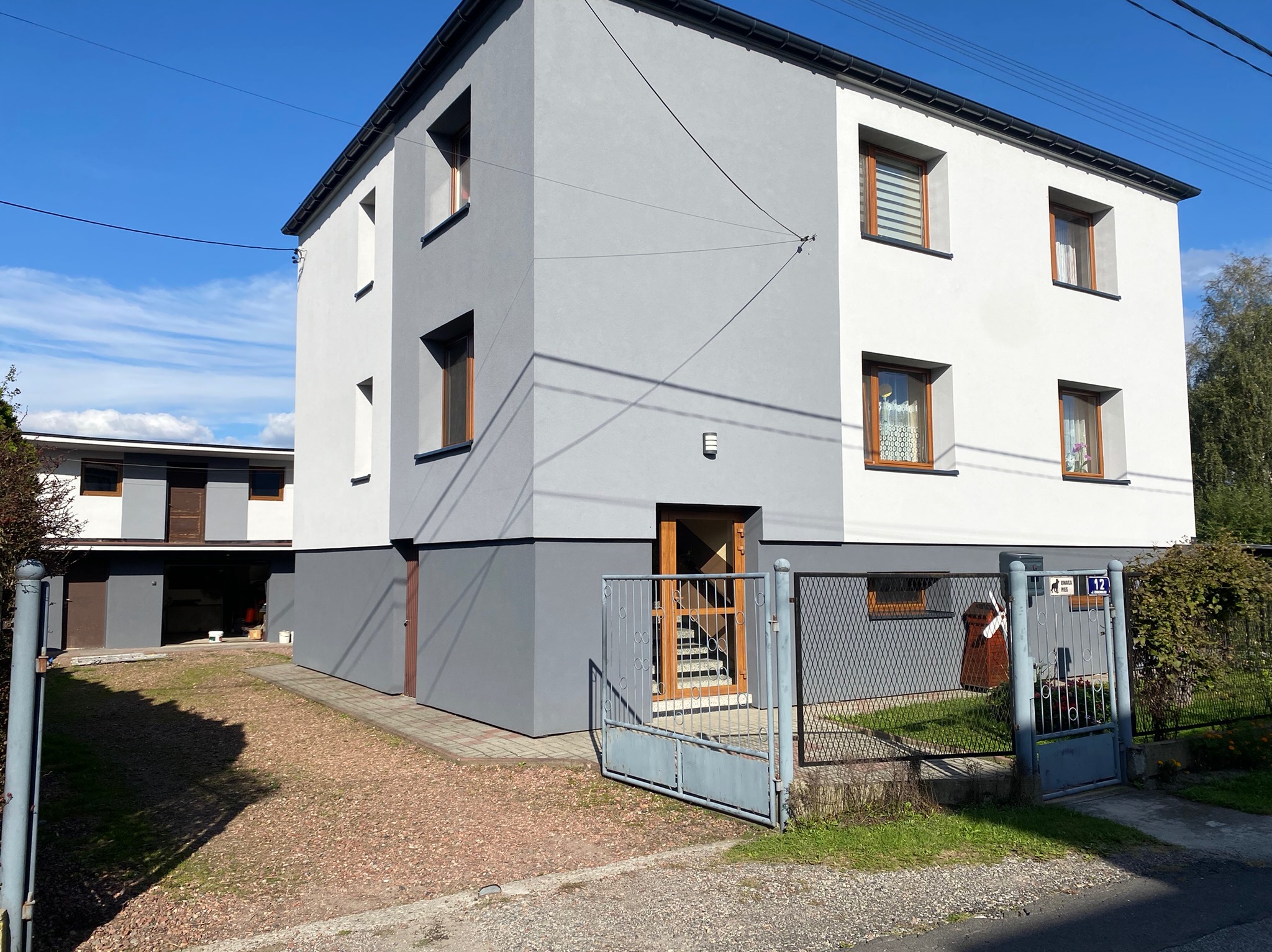 Dwukondygnacyjny dom z elewacją w odcieniach szarości i bieli, widoczny z zewnątrz przez otwartą, metalową furtkę i niski płot.