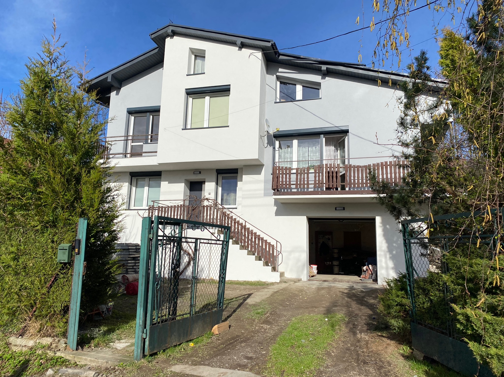 Dwukondygnacyjny dom jednorodzinny z jasną elewacją, brązowymi balustradami balkonowymi i schodami zewnętrznymi, widoczny przez otwartą, zieloną bramę wjazdową.