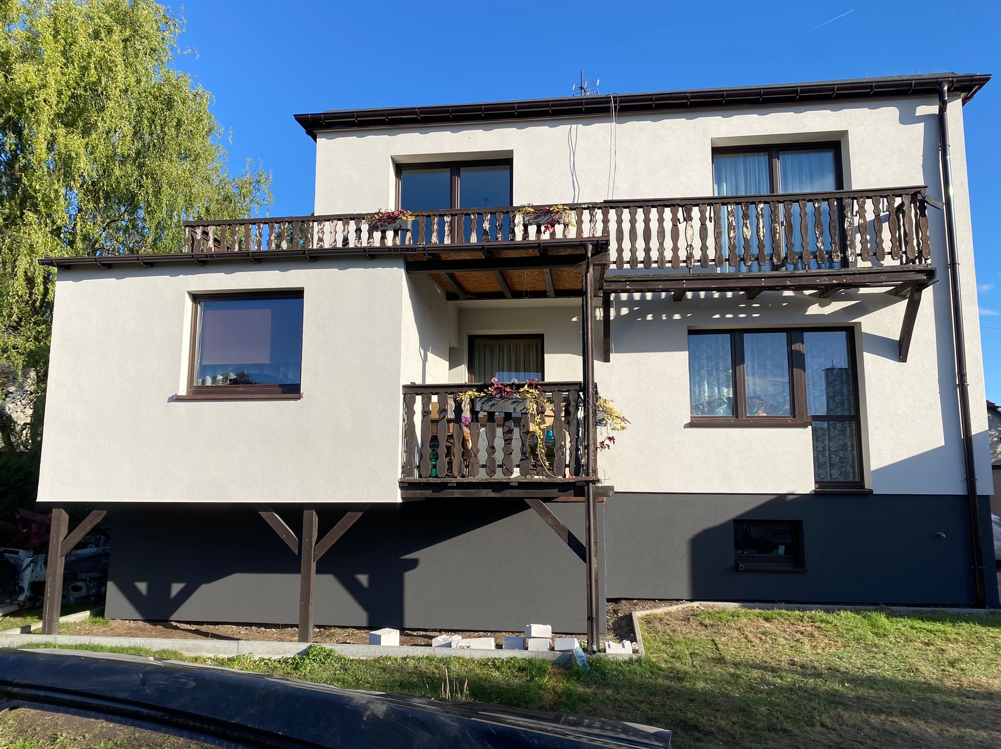 Dwukondygnacyjny dom jednorodzinny z balkonami w Bestwince, otynkowany na biało z ciemnoszarą dolną partią elewacji, drewniane balustrady balkonów z donicami kwiatów, błękitne niebo w tle.