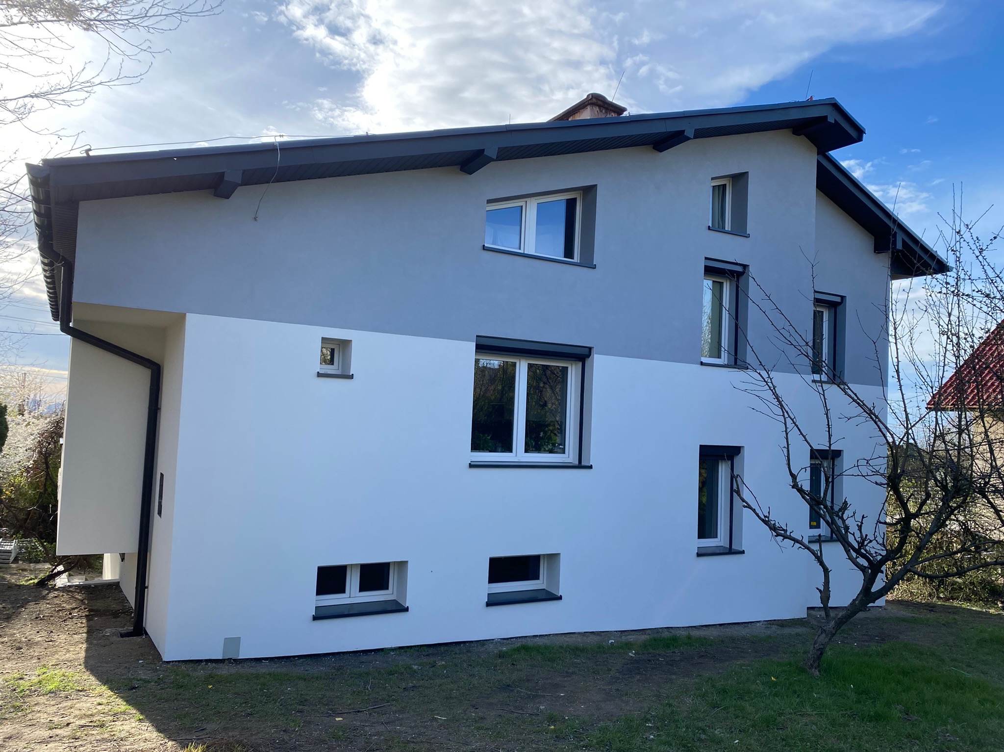 Dwukondygnacyjny dom po remoncie elewacji, z wyraźnym podziałem kolorystycznym: górna część szara, dolna biała, z ciemnymi obramowaniami okien i dachu.