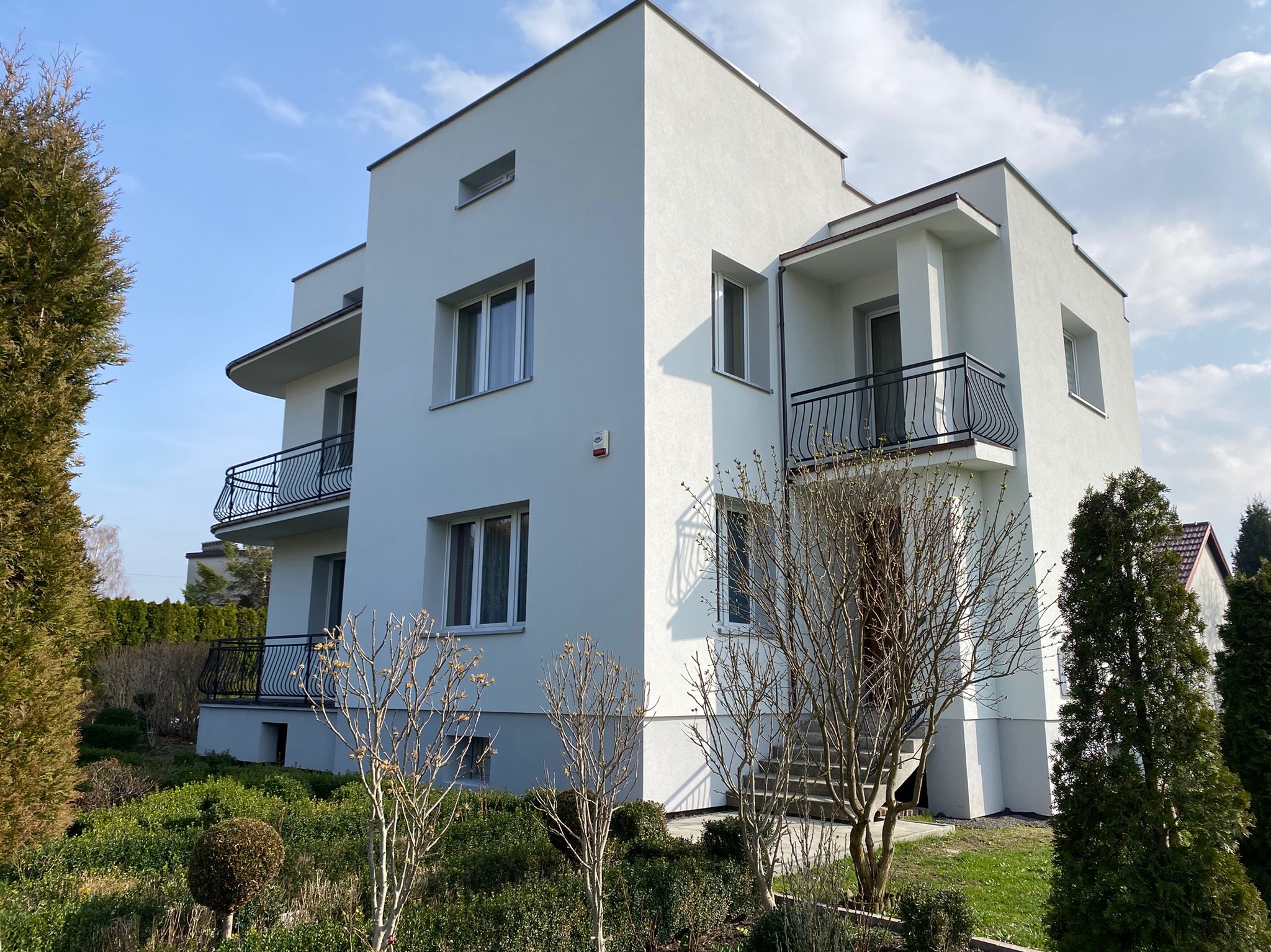 Nowoczesny, dwupiętrowy budynek mieszkalny z białymi elewacjami, balkonami z czarnymi balustradami i małymi oknami, otoczony zielenią i drzewami w słoneczny dzień.