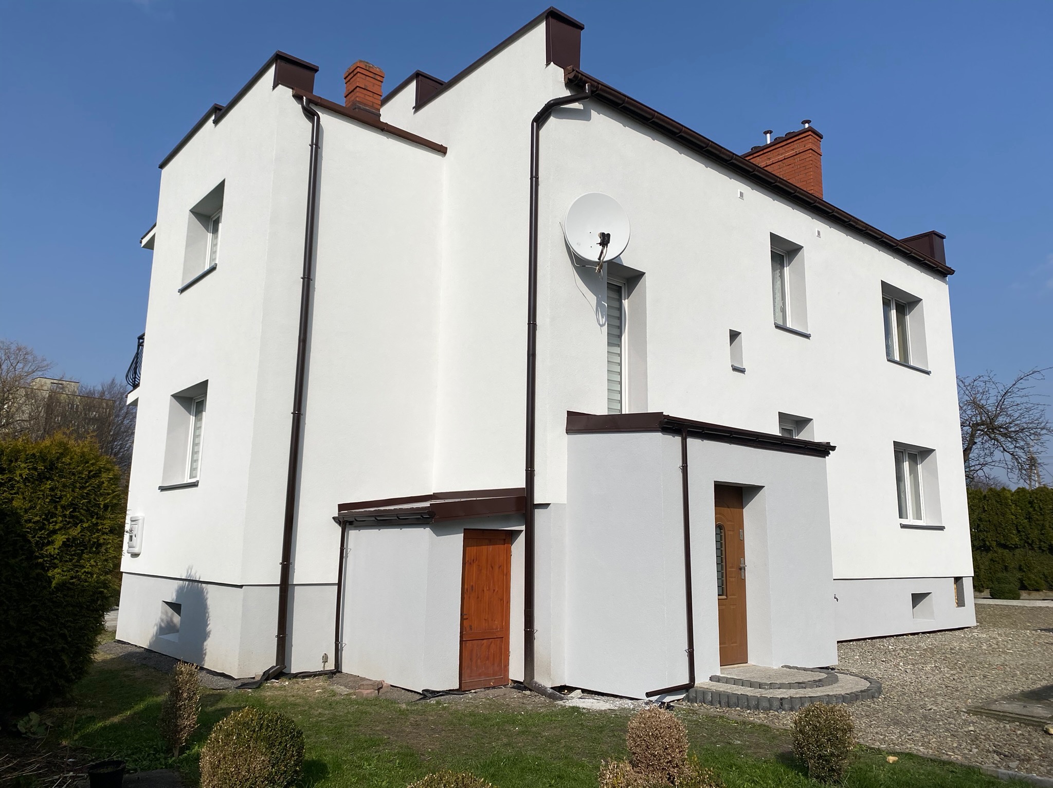 Trzykondygnacyjny dom jednorodzinny z białą elewacją, brązowymi elementami wykończeniowymi i anteną satelitarną, sfotografowany w słoneczny dzień.