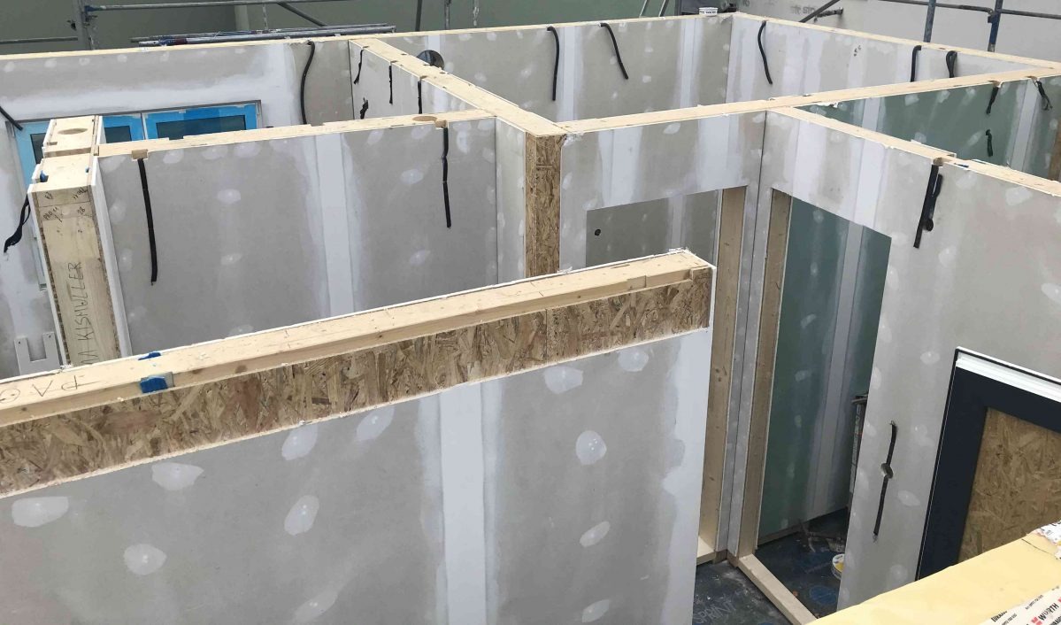 Wnętrze budynku w trakcie budowy, widoczne drewniane ramy konstrukcyjne i płyty gipsowo-kartonowe, z widocznymi przewodami elektrycznymi i otworami na okna i drzwi.