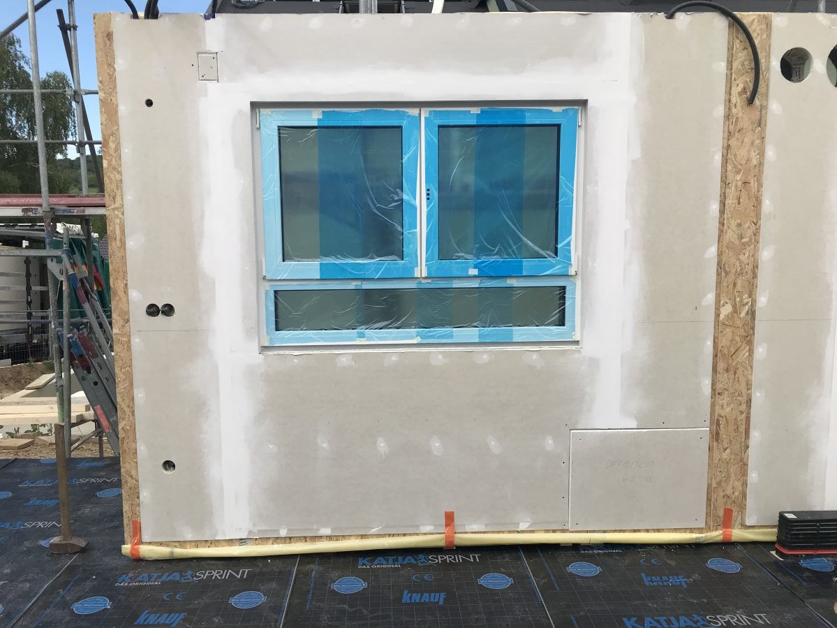 Wnętrze w trakcie budowy, widoczna ściana z oknem zabezpieczonym folią, wykończona płytami gipsowo-kartonowymi, z widocznymi łączeniami i przygotowaniem do dalszych prac wykończeniowych.