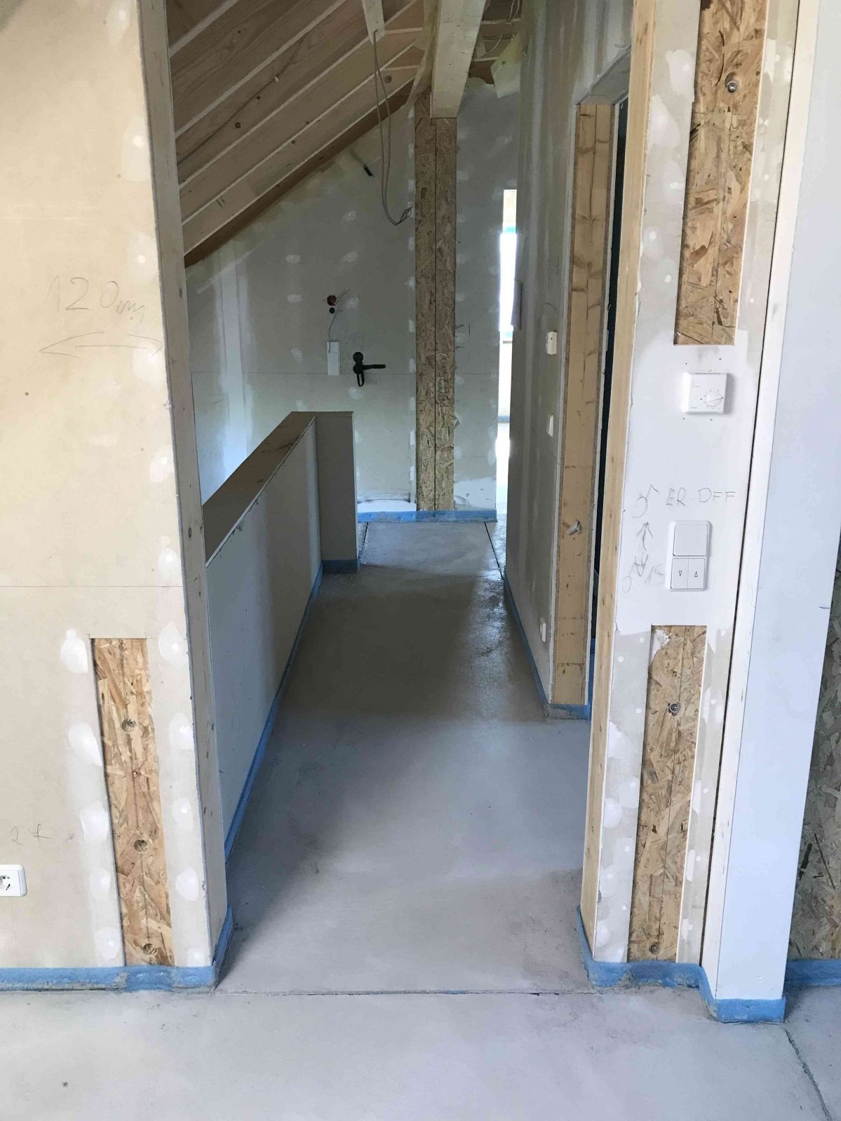 Wnętrze domu szkieletowego w trakcie budowy: widoczne surowe ściany z płyt gipsowo-kartonowych, drewniana konstrukcja szkieletowa, wylewka podłogowa, instalacja elektryczna i oznaczenia na ścianach.