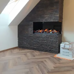 Wnętrze poddasza z oknem dachowym i dekoracyjnym kominkiem elektrycznym obłożonym kamieniem elewacyjnym, ułożona podłoga w jodełkę, widoczne kartony z okładziną.