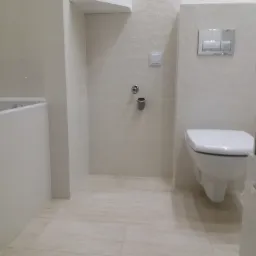 Jasna łazienka z kremowymi płytkami o wzorze plastra miodu, wisząca toaleta z chromowanym przyciskiem spłukiwania, zabudowana szafka nad toaletą i akcesoria łazienkowe.