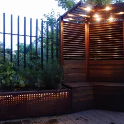 Nowoczesny kącik wypoczynkowy w ogrodzie z drewnianą altaną z lamelami, oświetleniem punktowym, metalowym ogrodzeniem i podświetlaną donicą z roślinami na drewnianym tarasie.