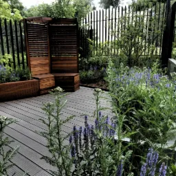 Nowoczesny taras kompozytowy z drewnianą zabudową, skrzynią na rośliny i nasadzeniami lawendy oraz krwawnika, ogrodzony czarnym metalowym płotem.