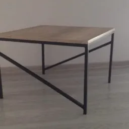 Kwadratowy stolik kawowy z blatem z jasnego drewna i czarną, metalową konstrukcją z ukośnymi wzmocnieniami, ustawiony na jasnej podłodze przy białej ścianie.