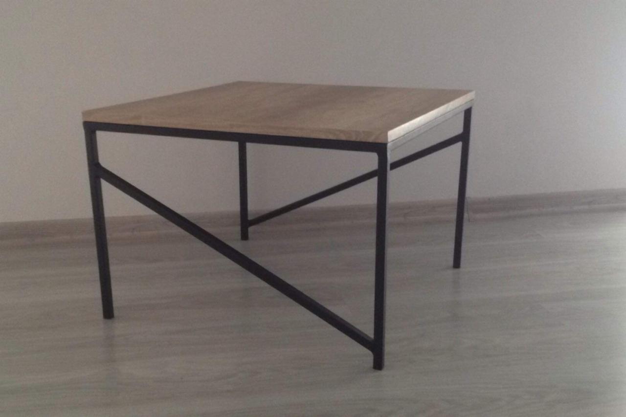 Kwadratowy stolik kawowy z blatem z jasnego drewna i czarną, metalową konstrukcją z ukośnymi wzmocnieniami, ustawiony na jasnej podłodze przy białej ścianie.