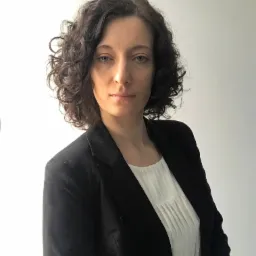 Portret kobiety z kręconymi włosami w czarnej marynarce i białej bluzce na tle białej ściany.