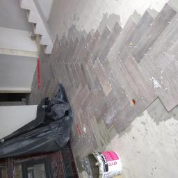 Glazurnik - Układanie płytek podłogowych w jodełkę, widoczne krzyżyki dystansowe, wiadro z klejem Ceresit CT 177, folia ochronna na podłodze i poziomica przy ścianie.