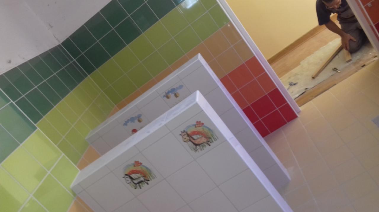 Kolorowa łazienka w trakcie wykańczania: ułożone płytki ścienne w odcieniach zieleni, żółci, pomarańczy i czerwieni, z motywami zwierząt na białych płytkach umywalki; glazurnik w tle, przygotowuje...
