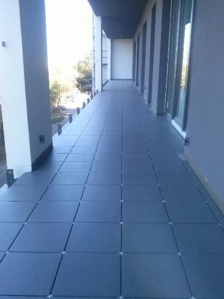 Długi balkon wyłożony ciemnymi, kwadratowymi płytkami z jasnymi krzyżykami dystansowymi, widok wzdłuż balkonu, szare ściany i filary.