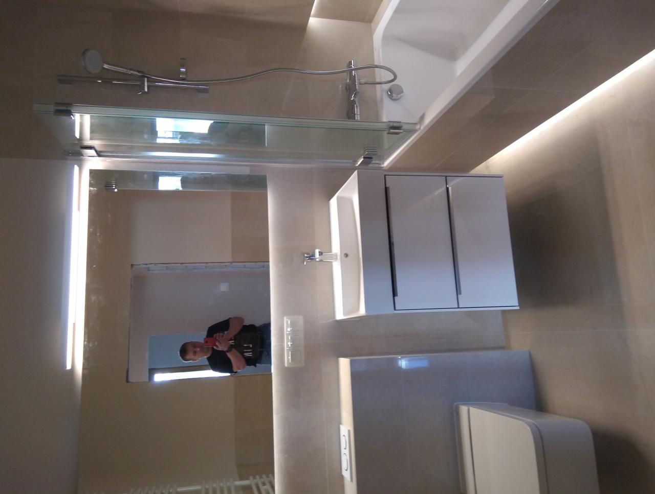 Nowoczesna łazienka z beżowymi płytkami, białą umywalką z szafką, wanną, toaletą i prysznicem ze szklanym panelem, odbicie osoby robiącej zdjęcie w lustrze nad umywalką.