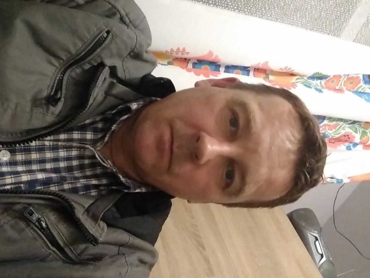 Portret mężczyzny w koszuli w kratę i kurtce na tle wzorzystej tapety i blatu. Ujęcie z bliska, obrócone o 90 stopni, z widocznym odkurzaczem.