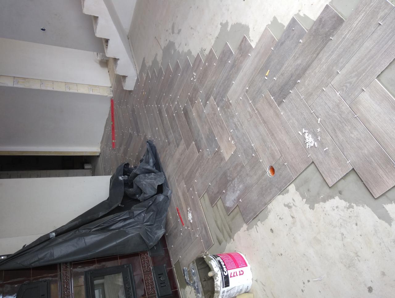 Układanie płytek podłogowych w jodełkę, widoczne krzyżyki dystansowe, wiadro z klejem Ceresit CT 177, folia ochronna na podłodze i poziomica przy ścianie.