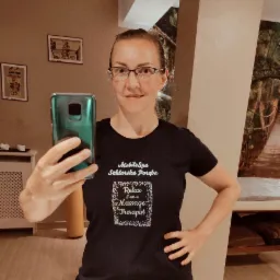 Kobieta w okularach robi selfie w gabinecie. Na koszulce napis 'MobileSpa Szklarska Poręba Relax I am a Massage Therapist'. W tle widoczny stół do masażu.