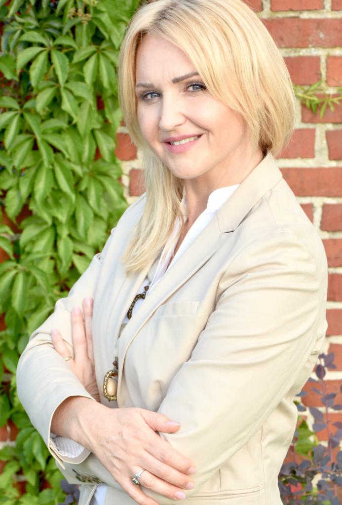 Uśmiechnięta blondynka w beżowym żakiecie, skrzyżowane ręce, na tle ceglanej ściany z bluszczem. Profesjonalny wizerunek agenta nieruchomości.