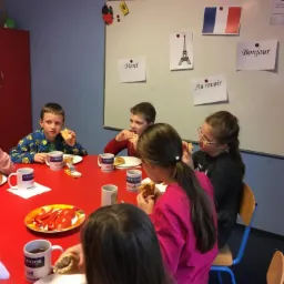 Grupa dzieci przy czerwonym stole je śniadanie; na ścianie tablica z francuską flagą, wieżą Eiffla i napisami 'Merci', 'Bonjour', 'Au revoir'.