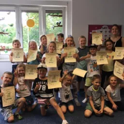 Grupa uśmiechniętych dzieci i nauczycielka trzymających dyplomy ukończenia kursu językowego w jasnej sali z dekoracjami okiennymi.