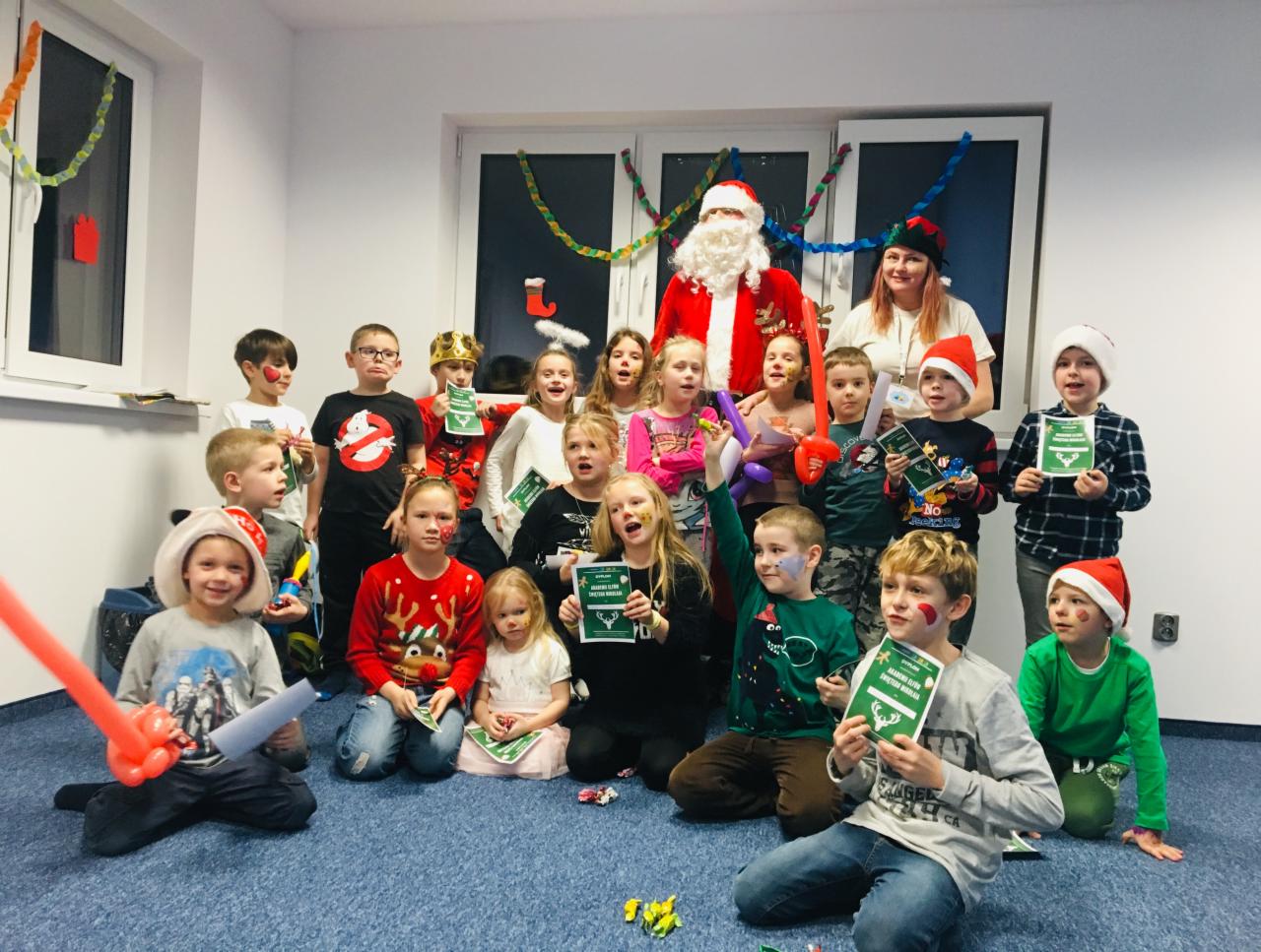 Grupa dzieci w różnym wieku, niektóre w świątecznych strojach i z pomalowanymi twarzami, pozuje z Mikołajem i kobietą w sali z dekoracjami świątecznymi, trzymając zielone dyplomy.