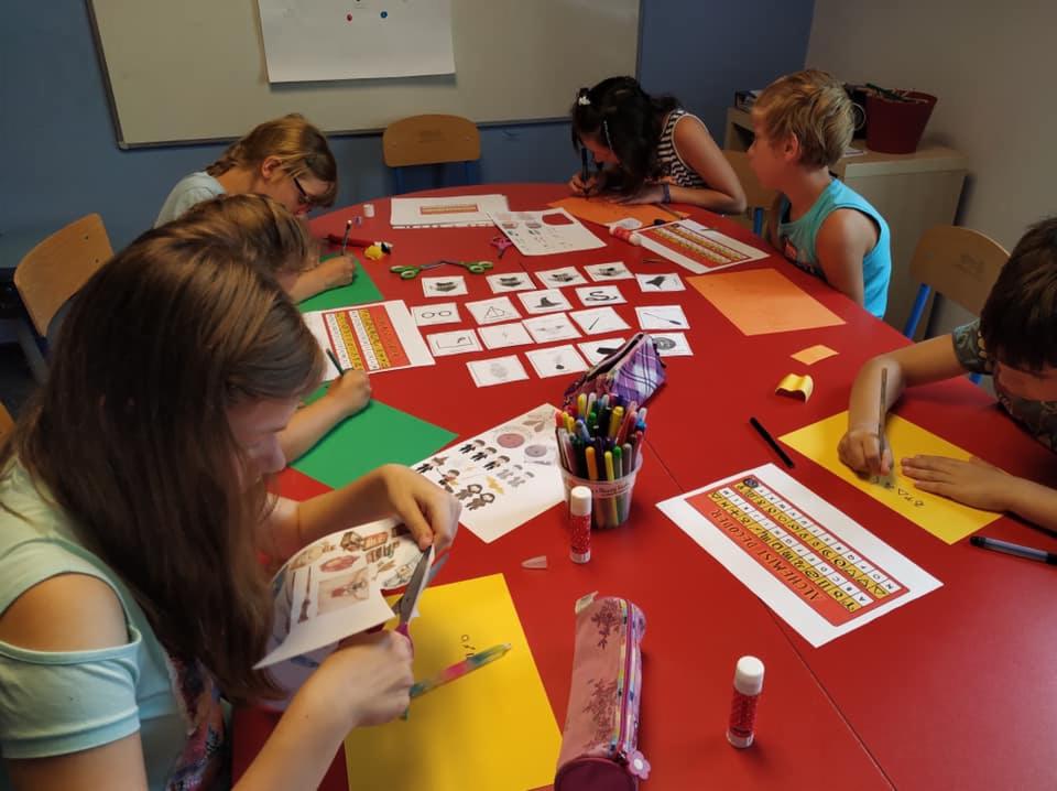 Grupa dzieci siedzi przy czerwonym stole, wykonując ćwiczenia z języka obcego, wycinając obrazki i pisząc na kolorowych kartkach, pomoce dydaktyczne, kleje i flamastry rozłożone na stole.