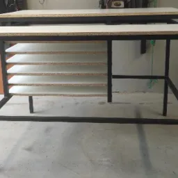 Dwa metalowe biurka z drewnianymi blatami i półkami, wykonane na zamówienie, widoczne w surowym wnętrzu, potencjalnie w warsztacie.