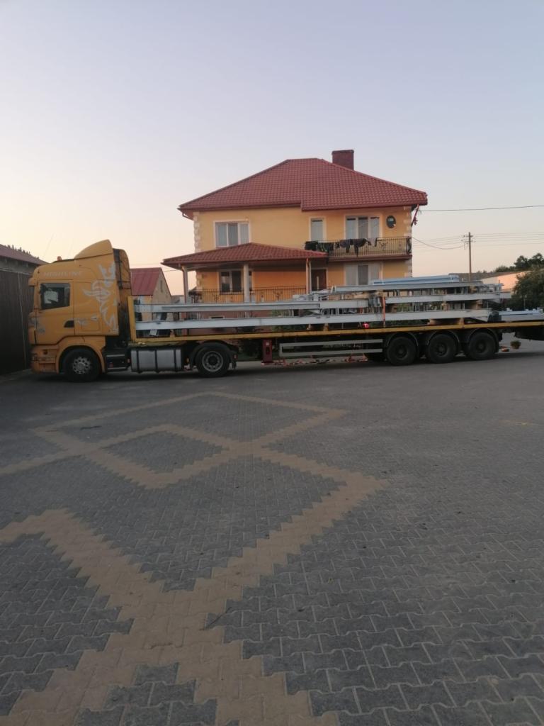 Żółta ciężarówka Scania z naczepą przewożąca stalowe elementy konstrukcyjne na tle dwupiętrowego domu z czerwonym dachem o zachodzie słońca.