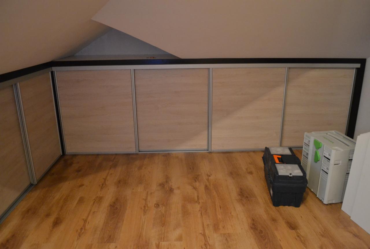 Zabudowa pod skosem z przesuwnymi drzwiami w kolorze jasnego drewna, skrzynka narzędziowa i białe opakowania po prawej stronie, podłoga drewniana.