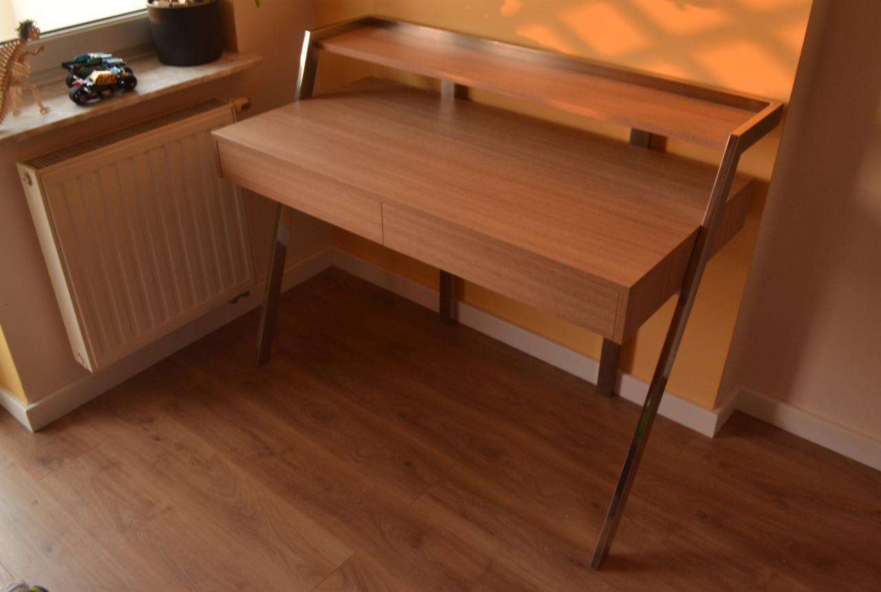 Nowoczesne biurko z drewna w ciepłym odcieniu, z metalowymi nogami, ustawione przy ścianie w pokoju z drewnianą podłogą i grzejnikiem pod oknem.