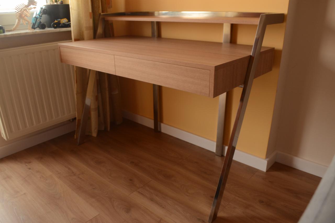 Nowoczesne biurko z blatem w kolorze jasnego drewna, metalowe nogi i górna półka, widok z boku na tle pomarańczowej ściany i drewnianej podłogi.