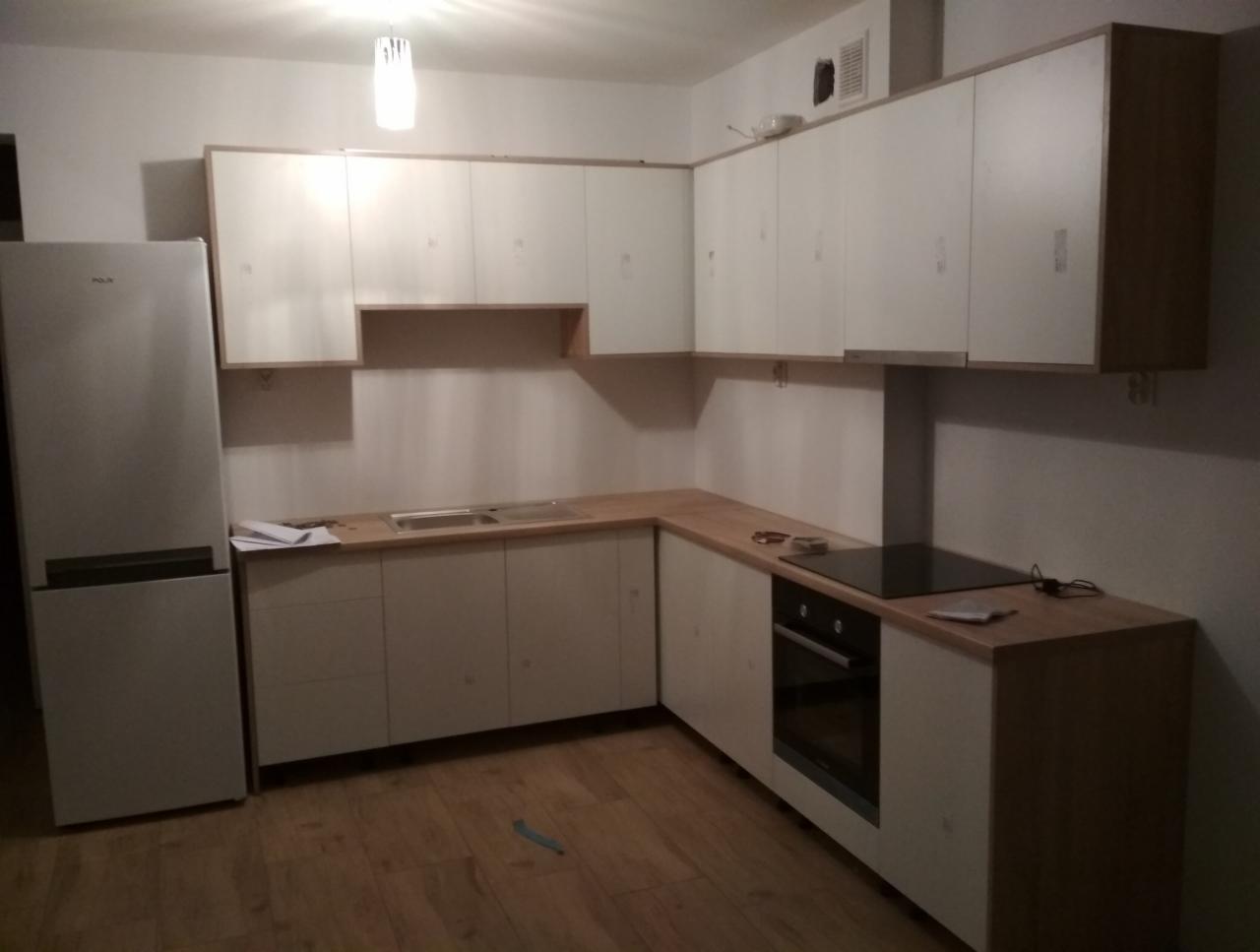 Jasna kuchnia z drewnianym blatem, białe fronty szafek, widoczna płyta indukcyjna i piekarnik, na blacie leżą dokumenty, obok lodówka.