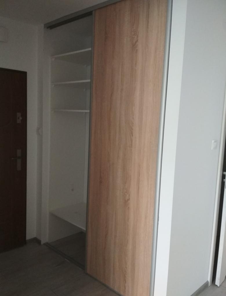 Wnętrze przedpokoju z zamontowaną szafą wnękową z przesuwnymi drzwiami o drewnianym wzorze, widoczne półki w środku, szara podłoga.