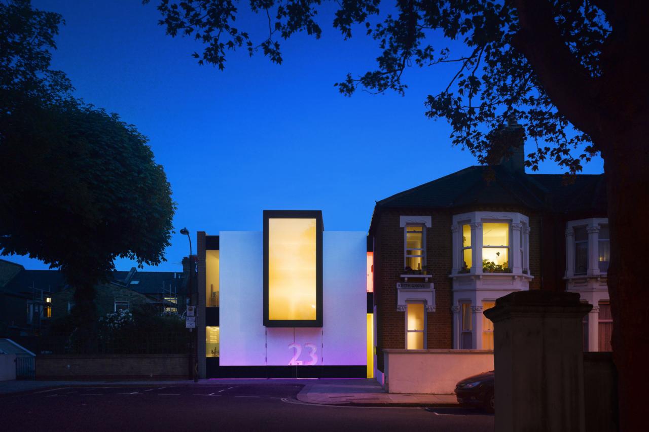 Nowoczesna elewacja domu z dużym oknem i podświetleniem numeru 23 w kolorze fioletowym, sfotografowana o zmierzchu, z widocznymi sąsiednimi budynkami i drzewami.