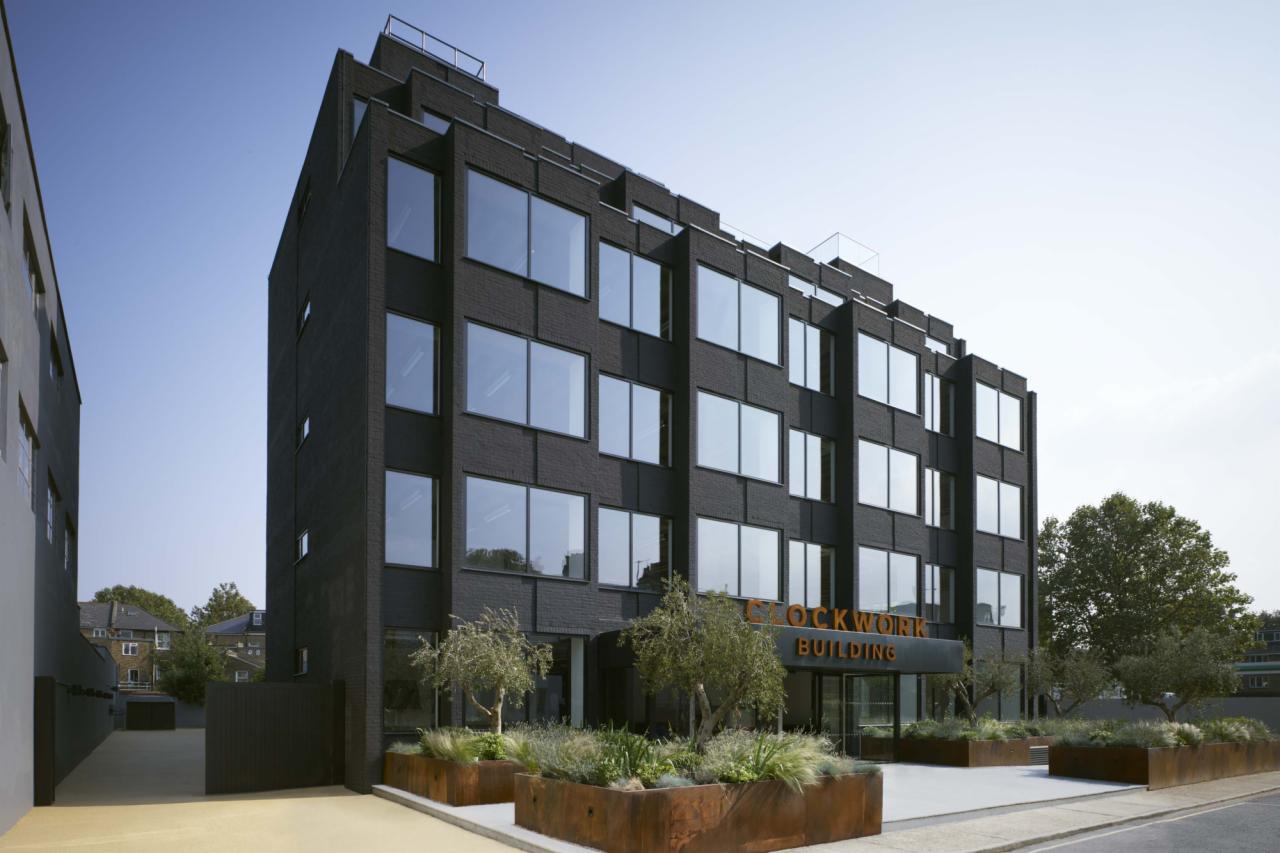 Czarny, modernistyczny budynek biurowy z dużymi oknami, nazwą 'CLOCKWORK BUILDING' nad wejściem i roślinnością w metalowych donicach.