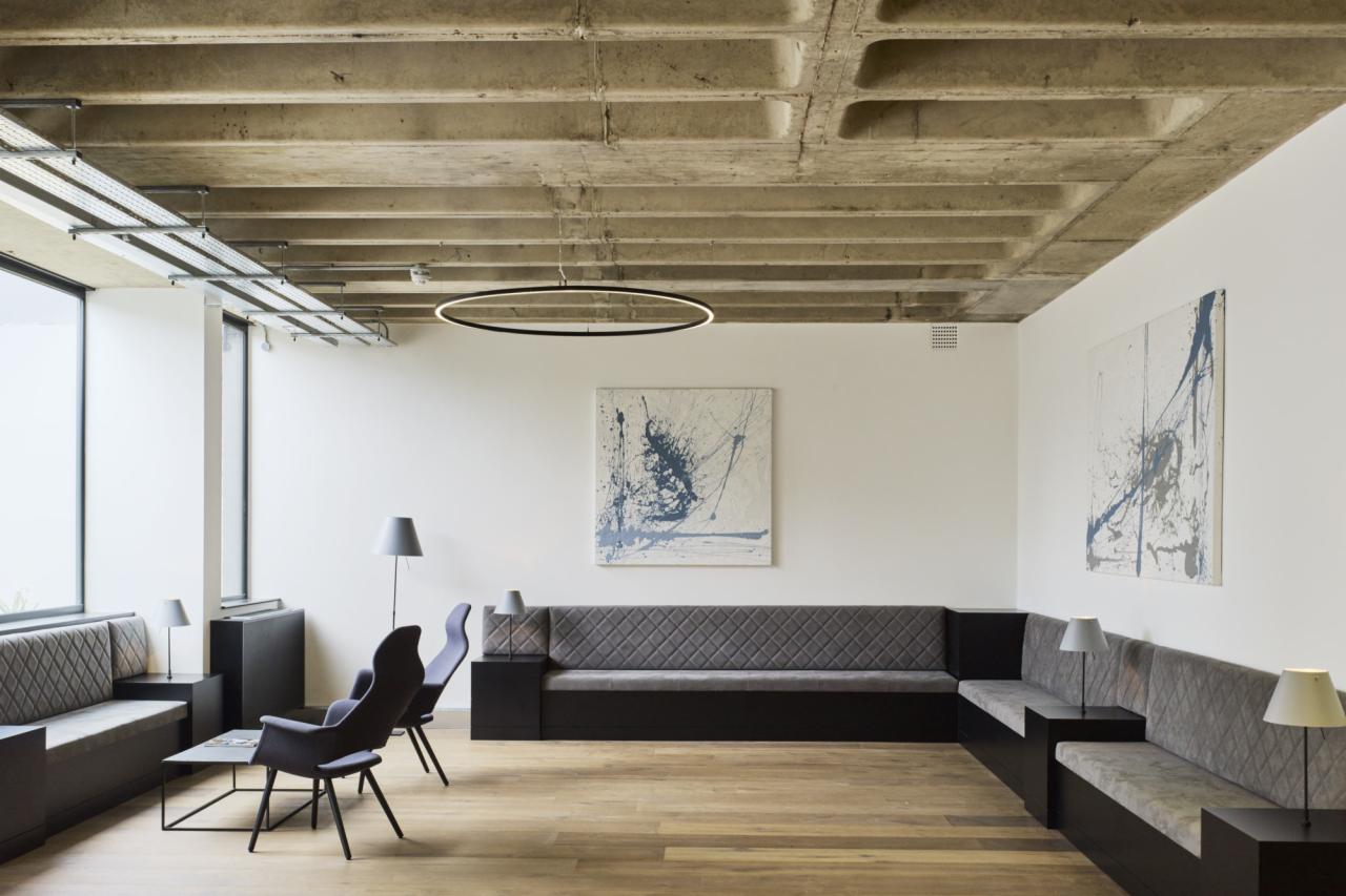 Nowoczesna przestrzeń biurowa z szarymi sofami w kształcie litery L, dwiema abstrakcyjnymi grafikami na ścianie i surowym betonowym sufitem z okrągłą lampą LED.