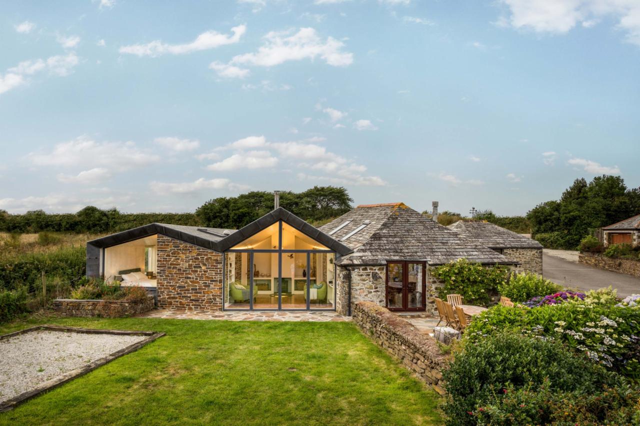 Nowoczesna bryła domu z kamienia, z przeszklonym frontem i widocznym wnętrzem, otoczona zielonym trawnikiem i kamiennym murem.