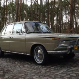 Beżowy, klasyczny samochód BMW w lesie, zaparkowany na brukowanej drodze, widok z przodu i boku, chromowane detale, białe opony.