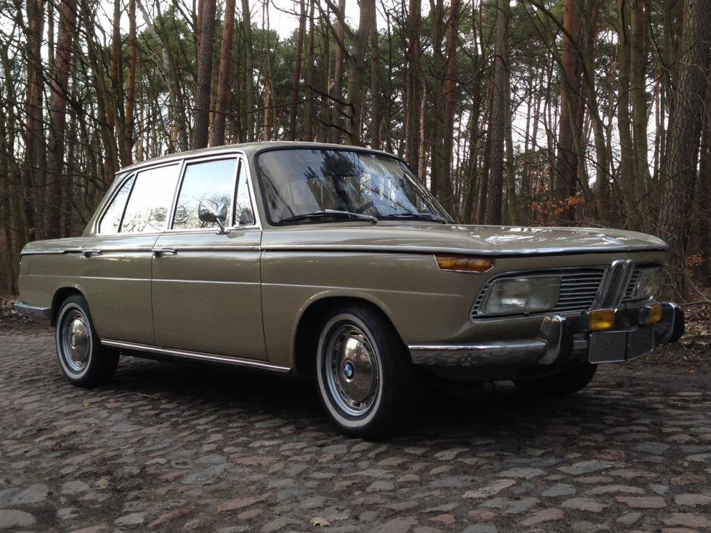 Beżowy, klasyczny samochód BMW w lesie, zaparkowany na brukowanej drodze, widok z przodu i boku, chromowane detale, białe opony.