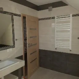 Wykończona łazienka na poddaszu z beżowo-szarymi płytkami, grzejnikiem drabinkowym, umywalką z szafką i lustrem w ramie, widoczne otwarte drzwi z drewnianą okleiną i dekoracyjnymi paskami.